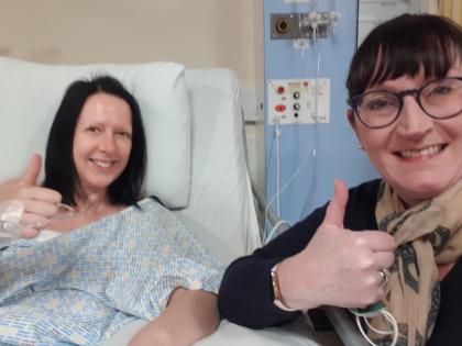 Gillian 2 days after her operation smiling in the hospital bed with a friend both giving a thumbs up